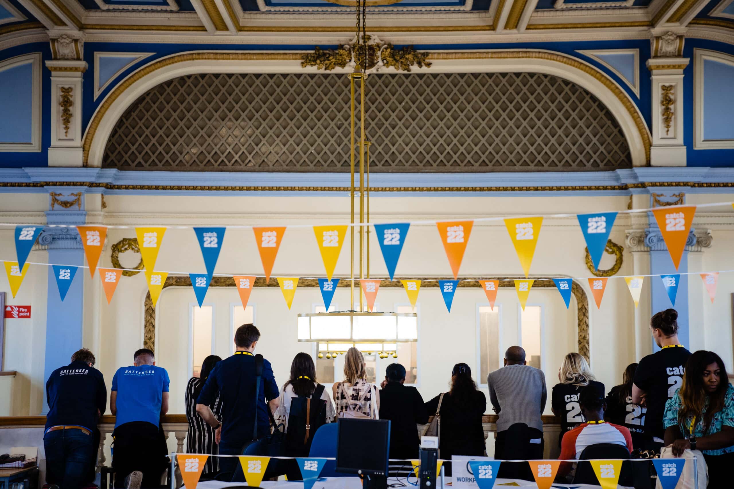 A group of people stand in front of a balcony listening to someone speak on the floor below. They all have their backs to the camera. Above them is Catch22-branded bunting.